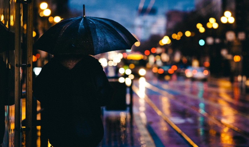 Ihmishahmo seisoo selkä kameraan päin sateenvarjon kanssa hämärässä kaupunkimaisemassa, autojen valot ja katulamput valaisevat maisemaa.